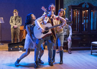 Autolycus wearing flower embroidered tan jacket, sideburns and red glasses with clip-on shades plays an acoustic guitar as Mopsa in short white dress strokes his leg and Dorca in knee-lenth jeans shorts hugs his shoulders. Polixenes disguised with beard and brown volksmarch suit stands in the background, Camilo sits on a chair next to the empty china cabinet in the back right