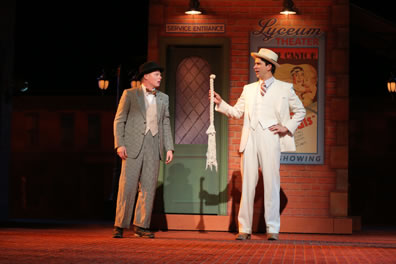 Dromio in green-pinstriped suit looks at Antipholus in white three-piece suit holding a short rope with a knot at one end. Behind is a wall and door with a poster for the Lyceum