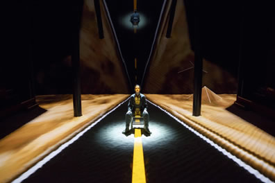 Hathaway wearing a flightsuit sitting in a chair in the middle of a road, the yellow strip passing right through her, with golden sand on either side and a pyramid in the background