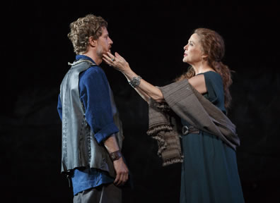 Goneril on the right strokes Edmund's beard with her fingertips; she is wearing a blue dress with a gray scarf draped over her arms and around her waist, he is in blue shirt, gray pants and gray leather vest.