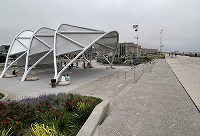 The amphitheater from the side, clearly showing its silver-metal three nested awnings that comprise the roof. To the right is the Rockaway Beach Boardwalk 