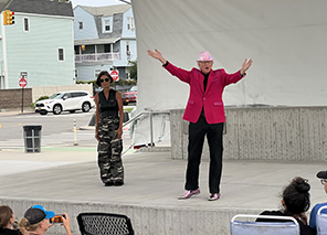 Theseus in red jacket, black pants, and pink hat and arms outspread, next to him is Hippolyta in camoflage pants and dark vest on a concreet stage with houses and a street intersection in. the background, the heads of people in lawn chairs in the foreground.