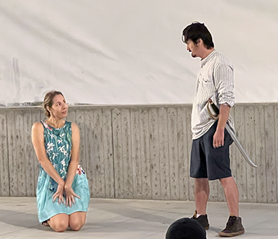 Helena wearing an aqua sun dress with white blooms, kneels on the stage with her hands obediently pleading with Demetrius standing across from her wearing a kacki shirt, olive shorts, and a sword in his belt. 