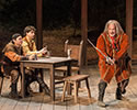 Production photo of Falstaff weilding a bent, hacked swoard as Poins and Hal sit at a table watching.