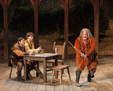 Production photo of Falstaff weilding a bent, hacked swoard as Poins and Hal sit at a table watching.