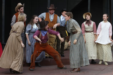 Several people in old West clothing watch two women face off as Pompey in the middle, wearing purple shirt and red pants, stands between the combatants backing them away from each other