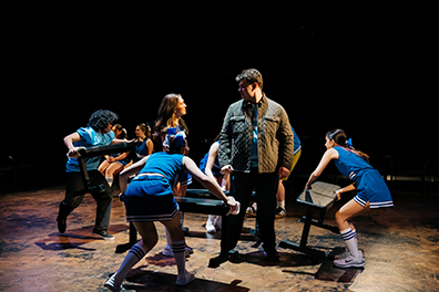 SIG-This_Ends_In_Blood-Mac&Beth-4 Beth wearing a blue cheerleader uniform looks up at Mac wearing an olive flack jacket over a blue football jersey and black pants, standing opposite each other as three cheerleaders and a football player carry and place benches for the next scene and two other cheerleaders sit side-by-side in the background in position to speak their first lines.