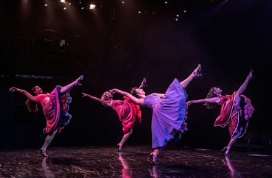 Four women in '50s frilly hispanic pary dresses and high heels, standing on left legs, right legs kicking high, leaning back so spines are parallel to the stage, hands extended back and away.