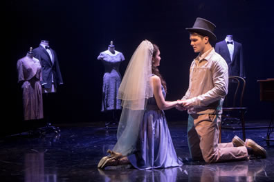Maria and Tony kneel on the stage and holding hands,  she in simple dress and wearing a wedding veil, he in brown work shirt and pants wearing a black tophat. In the background are maniquins with formal suits and nice dresses