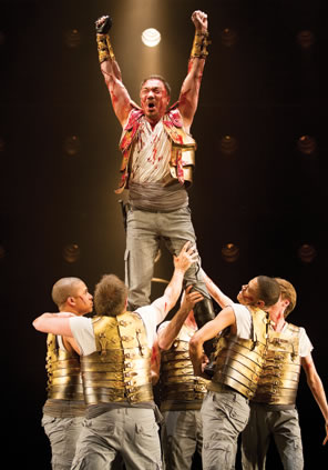 Coriolanus, arms upraised, bload on his gold armor and uniform, is lifted high above by the other soldiers.