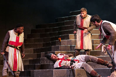 Three soldiers wearing Saint George smocks over chain-mail armor stand around another soldier lying on the stone steps with an arrow sticking into his shoulder, blood on the smock, his hand gripping at his breast, a sword in his other hand. 