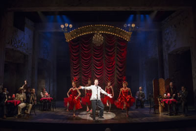 Lucio in white dinner jacket at a microphone and in spotlight with four burlesque dancers behind him in red in front of a red-curtained music hall stage and red tableclothed cabaret tables on either side.