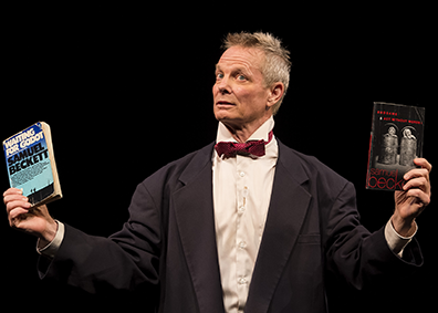 Bill Irwin, wearing a dinner  jacket, white shirt, and red bowtie, holds up in his right hand a well-read paper back edition of "Waiting for Godot" and in his left hand a paperback edition of "End Games" and "Acting Without Words."