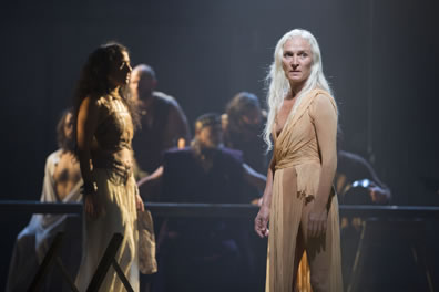 The nameless woman with long white hair and wearing a loose-fitting plain tan dress with young Salome in white pleated skirt and leather corset crosses behind; at a table in the background is a tableaux of men.