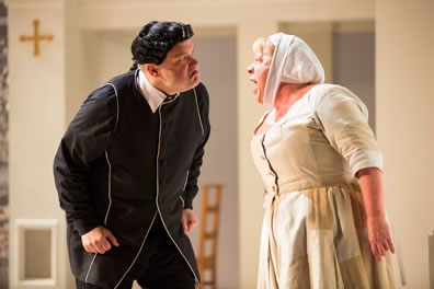 Orgon in a black frock and black wig with curls on the side, hands clenched at his waist, leans in toward Dorine in a simple maids dress and head scarf, shouting back.