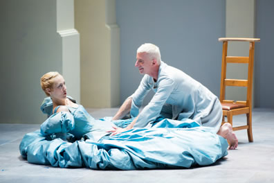 Tartuffe in pale blue smock and on his knees leans over Elmire in a blue ball gown, leaning back on the floor, hands propping her up, and a plain wood chair is behind Tartuffe