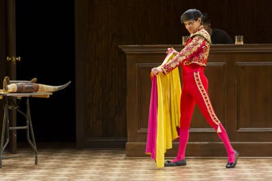 Romero is poised, right leg straight, left leg back, wearing jewelled jacket and red tight matador pants and black matador hat, yellow backed red cape spread in his hands, Before him is a table with bull horns attached to the front.