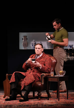Harry sitting in chair sipping from a tea cup he holds in his right hand over a saucer in his left, he's wearing a rust-patterned robe over a shirt and tie and black pants. Behind him, Bill in tight green pull-over shirt and checkered pants pours tea in his cup; he's standing next to a bar cart with crystal and bottles on top, and a line of Chinese porcelain in a glass case behind him. A Persian rug is on the bloor, and a wood end table and magazine rack are on either side of Harry's chair.