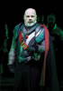 Wallenstein in renaissance armor, red and black cape and red sash facing the audience with ghostly images in the background.