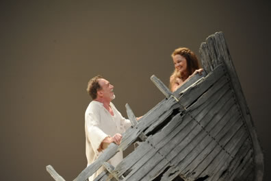Miranda in the bow of a wrecked boat looks back, smiling, at Prospero behind her in the bow and wearing a ragged white shirt.