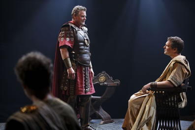 Antony stands in red skirt and cape with leather breastplate and decorative armor hanging down the sleeve and skirt and leather boots facing Octvius in white senator's toga sitting in a wood X-shaped chair with curved arms and legs, The back of somebody's head is in the left of the picture.
