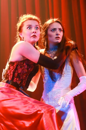 Helena in red dress and long black gloves and Hermia in white dress, long white gloves and jeweled necklace clasp each other with alarmed expressions.