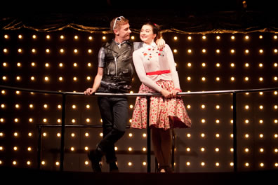 Claudio in leather fest and black jeans, sunglasses pushed up on top of his red head, standing cross legged, has his right hand on the rail and left hand draped around Hero's shoulder; she's wearing a white sweater with embroidered flowers, red polka dot skirt, and red belt; she's looking coyly at Claudio. Background of white lightbulbs