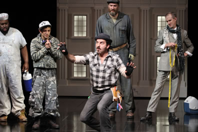 Bottom in checkered shirt and tool belt with his fingerless gloved hands as claws: watching behind him are Snout in white overalls and carrying a thermos, Flute in camouflag shirt and baggy pants with baseball cap, tall Snug in gray coveralls, and Starveling in striped suite, black scarf with white pattern around his neck, and holding a yellow tape rule. Lunch chest is on the floor behind, and the facade of a white building with pillars in the background.