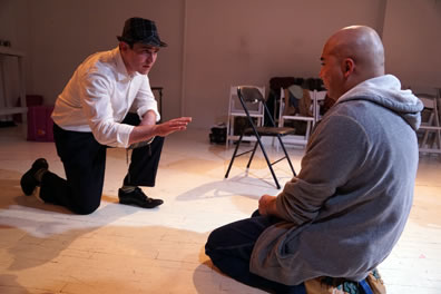 The Duke on one knee with white shirt, black pants and pork pie hat holds a Bible in his left hand and holds his right hand out toward Claudio in gray hoodie and black pants kneeling on the floor 
