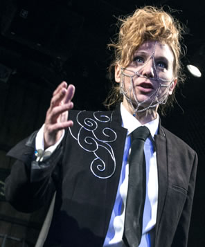 Poseidon in black suit jacket with silver scrolls on the right breast, white shirt, and black tie with necklace chain draped across her cheeks, down her forehead on either side of her nose, and dangling from her chin. 