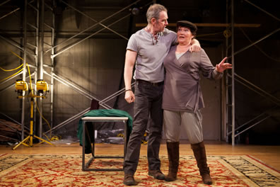 Gore with his arm tight around Packer's neck, she in tunic, dogcap and boots in a space with an oriental rug on the floor, a metal table behind the actors, lights, trash, and scaffolding against the wall