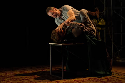 The two lying on the table, Gore on top with hand gripping her face as he screams at her, she fighting back, gripping his arm and kicking her leg up.