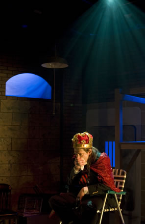 King John wearing a formal red and gold crown, red velvet cape and business suit sits on a green and whit striped lawn chair, leaning forward with hand over mouth, and a single spotlight shines down from the ceiling
