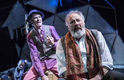 Lear in a white shirt, gray work pants, and a multi-color tapestry scarf with fringe around his shoulders; behind him the Fool in purple shirt, pink pants, gray vest, and blue hat talks, a finger held up; in the background, a servant in gray scarf sleeps