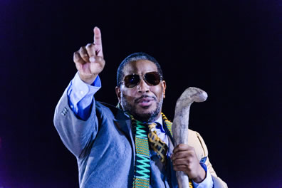 Oedipus in blue three-piece suit with yellow and blue diamond tie, sunglasses,an African scarf oer his shoulders, a wood crookstaff in his left hand while he raises the index finger of his right hand.