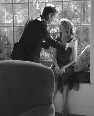 Benedick with his hands on Beatrice's shoulders as she sits on a window seat, trees visible through the window, pillow to one side of the seat, and the back of a plush chair in the foreground