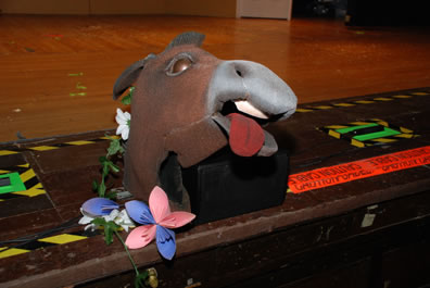 An ass's head, with tongue sticking out to one side, sits on the edge of a stage with construction paper flowers alongside.