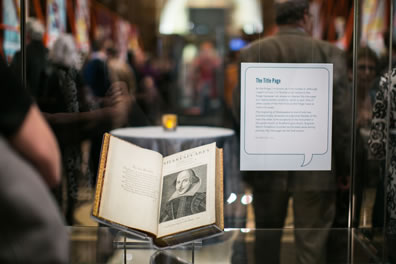 The First Folio, opened to the title page, in a glass case through which we can see a crowd of people in the exhibit, and a descriptive sign on the case titled "The Title Page."