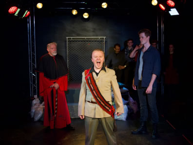 King John in tan uniform jacket and olive pants and a red and black sash across his chest stands front and center yelling; behin him to his right, the Cardinal is in red cardinal robes with black stole; to John's left King Philip in blue jeans, blue shirt and blue tee. The platform with bordered by chain link fences is at the back of the stage, sandbags at the base, and other cast members lined up against the backstage wall.