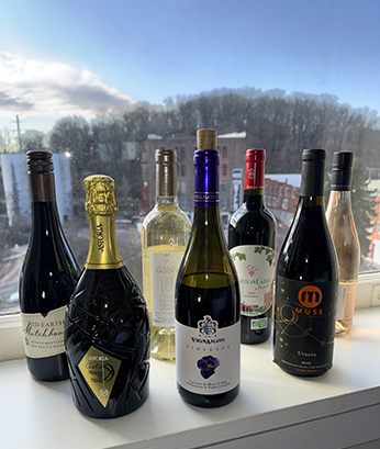 ASC-Twelfth_Night-32-261 Seven bottles of wine stand on a window sill with windery scene of buildings and a hill of bare trees through the window.
