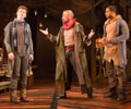 The Player with arms out and legs in a collosus stance talks with Guildenstern to his right, who is looking up and away, while Rosencrants on The Player's left looks at him. In the background is a stuffed chair behind Guildenstern and a attic ladder behind Rosencrantz and various junk. 