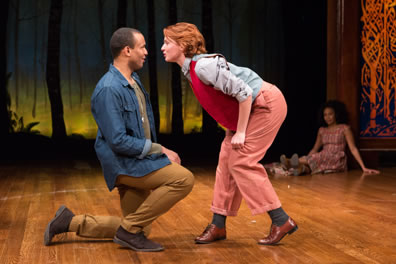 Orlando in blue denim shirt jacket, olive green long-sleeve tee, brown slacks and brown shoes is on one knee as Rosalind in her Ganymede disguse of red vest, denim shirt, pink slacks and brown patent leather shoes talks in his face. Celia, in a plaid country dress, sits on the stage in the background.