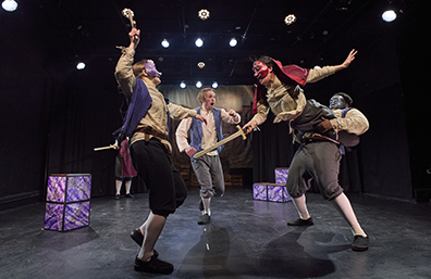 Tybalt, lifted in the air being held by Benvolio at his waist, sword fights with Mercutio, as Romeo runs towrd them. Marbelated segments of the chest stacked as part of the set. 