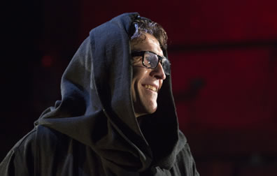 The Duke in a friars robe and hood, with dark-framed glasses and smiling off to the side.