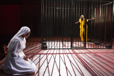 Isabella in nun's white habit on her knees, Claudio wearing yellow prison shirt and pants inside a jail cell, the bars casting shadows on the stage floor around the kneeling Isabella.