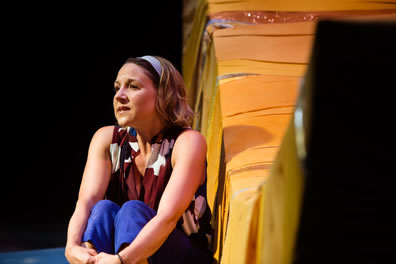 Jenn, in multi-colord tunic and blue pants, sits with her hands clasped around her shins with her bent legs pulled up to her body. Next to her is the bottom of the crescent moon.