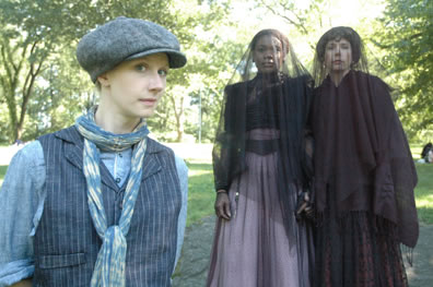 Viola in striped vest, scarfe and dog cap stands in front of a vieled Oliva and Maria, both in formal gowns.