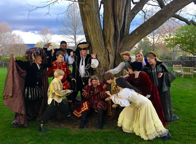 Around a tree in a park, various men and women in Elizabethan costumes with claws upraised close in on Falstaff sitting at the base of the tree.