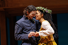 STC-CST_HAMNET William in a blue Elizabethan patterned vest jacket and shirt snuggles and holds hands with Agnes, wearing a white shirt and brown patterned dress and a crown of flowers.