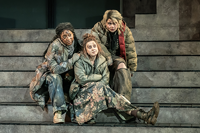 The three witches huddle on the steps to the bunker watching action toward the camera. They are wearing a colorful collection of baggy clothes and oversize coats, 